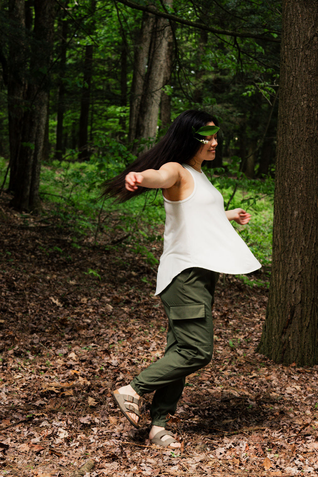 Person in a white sleeveless top and green pants running through a forest.