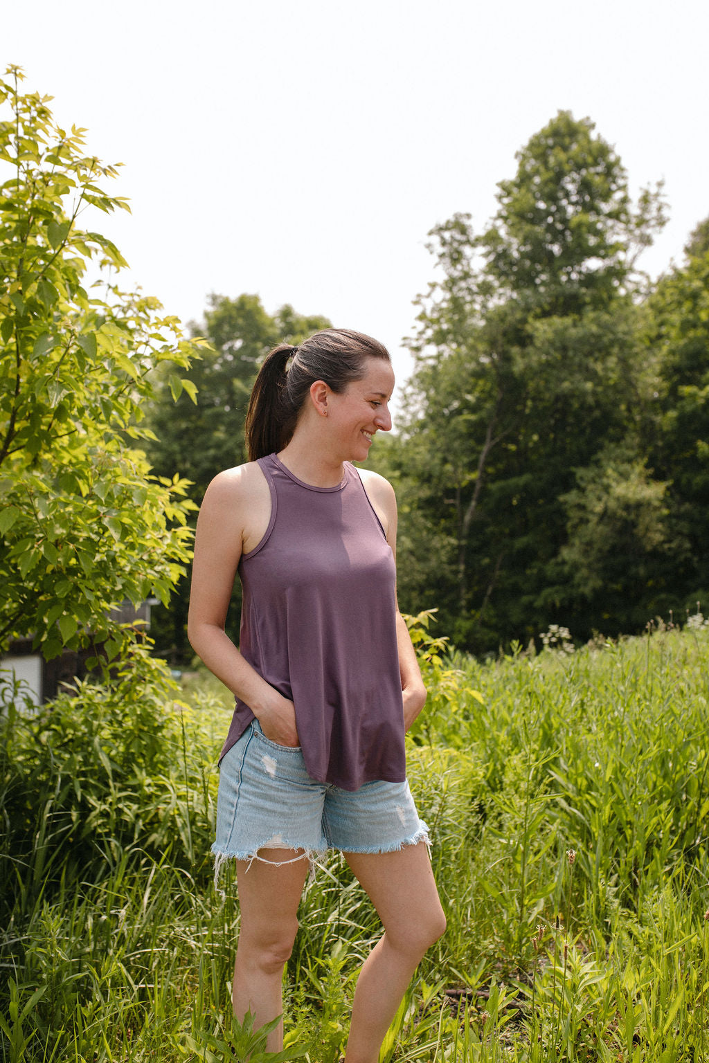 Woman wearing a lavender top standing in a grassy field with trees in the background