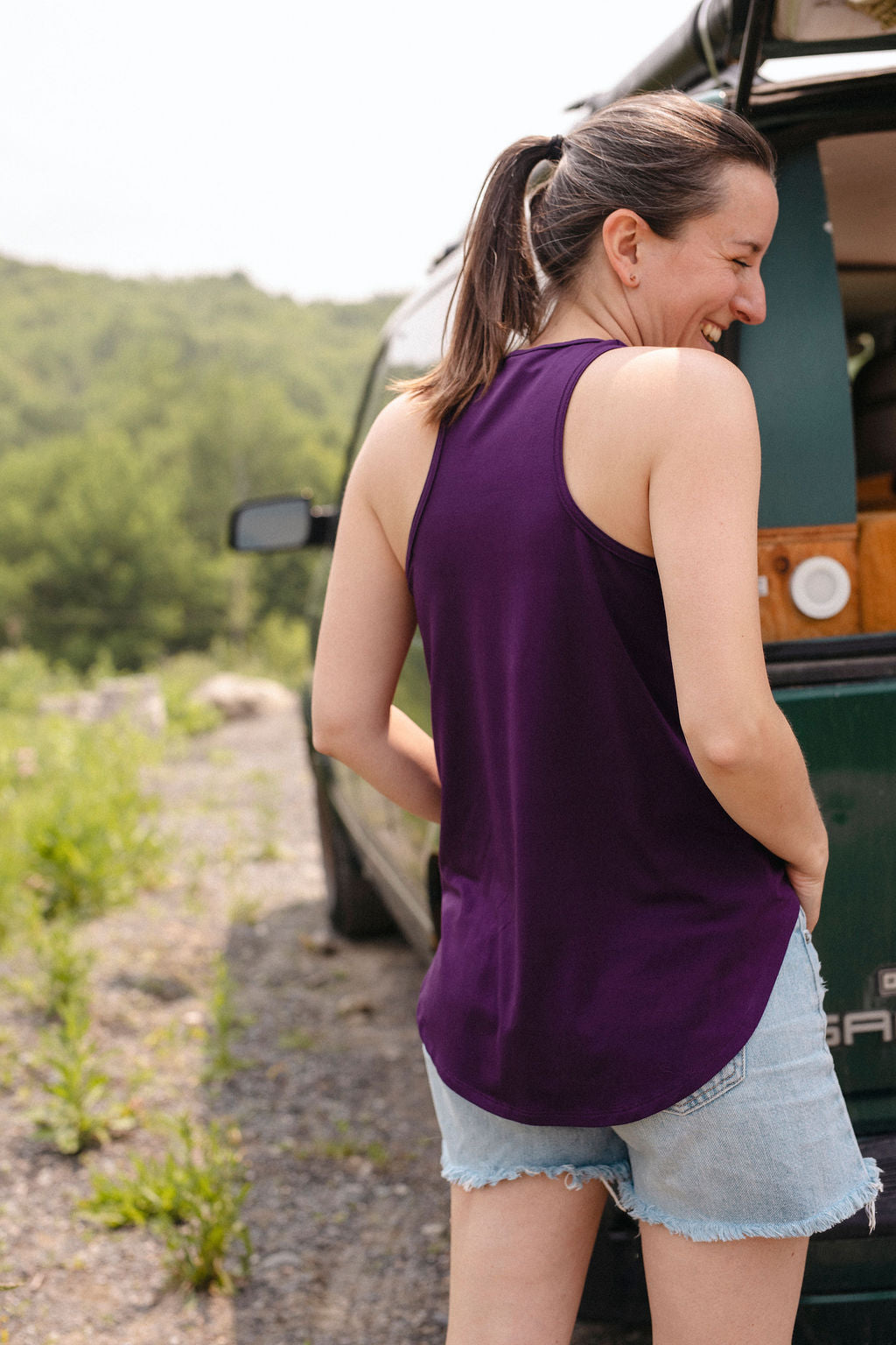 Woman in a purple tank top and denim shorts standing next to a green vehicle in a natural setting.