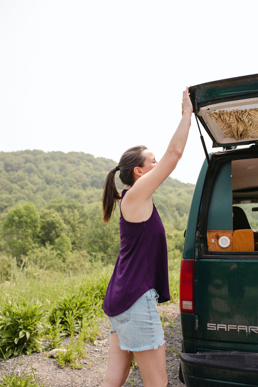 Woman wearing a plum top reaching into the open trunk of a green van with a scenic background