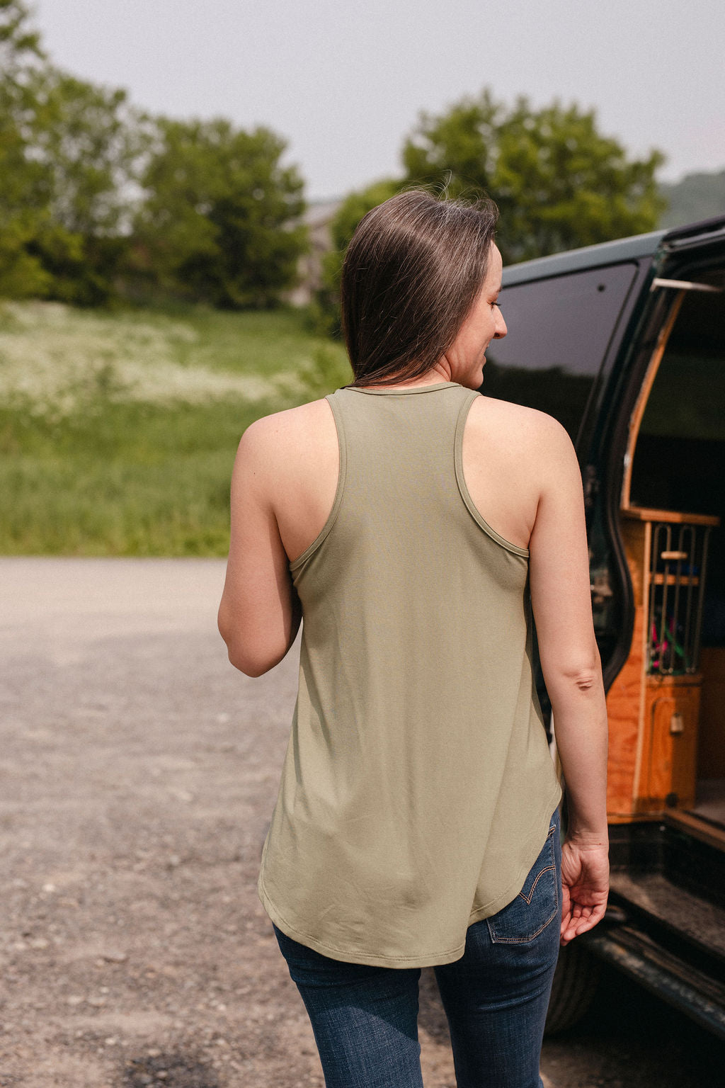 Back view of a woman wearing a sage green tank top, standing next to a van with nature in the background