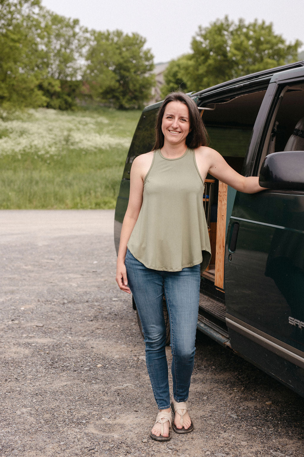 Woman wearing a sage tank top standing next to a green SUV in an outdoor setting with greenery.