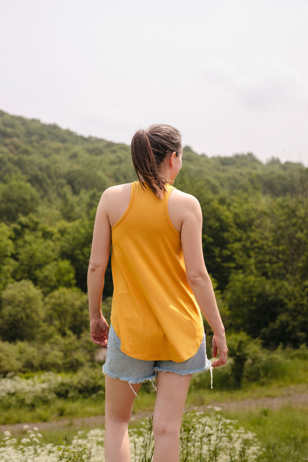 A woman wearing a yellow tank top and denim shorts, viewed from behind, standing in a forested area