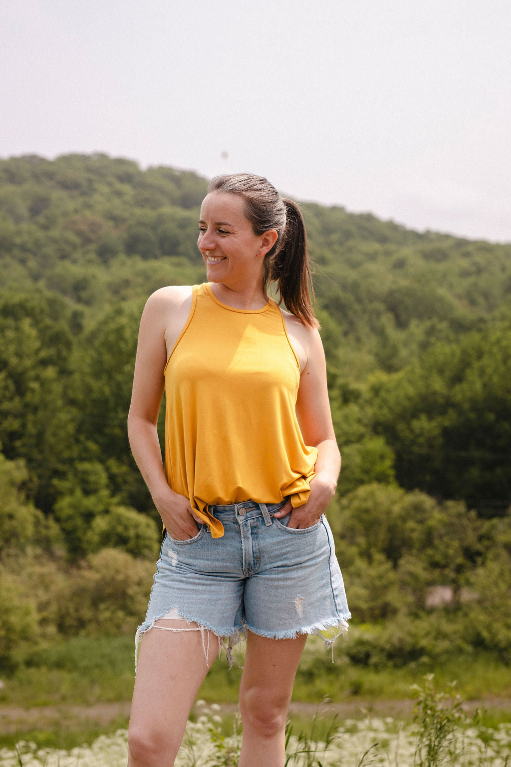 Woman in a yellow tank top and denim shorts standing in a field with trees in the background