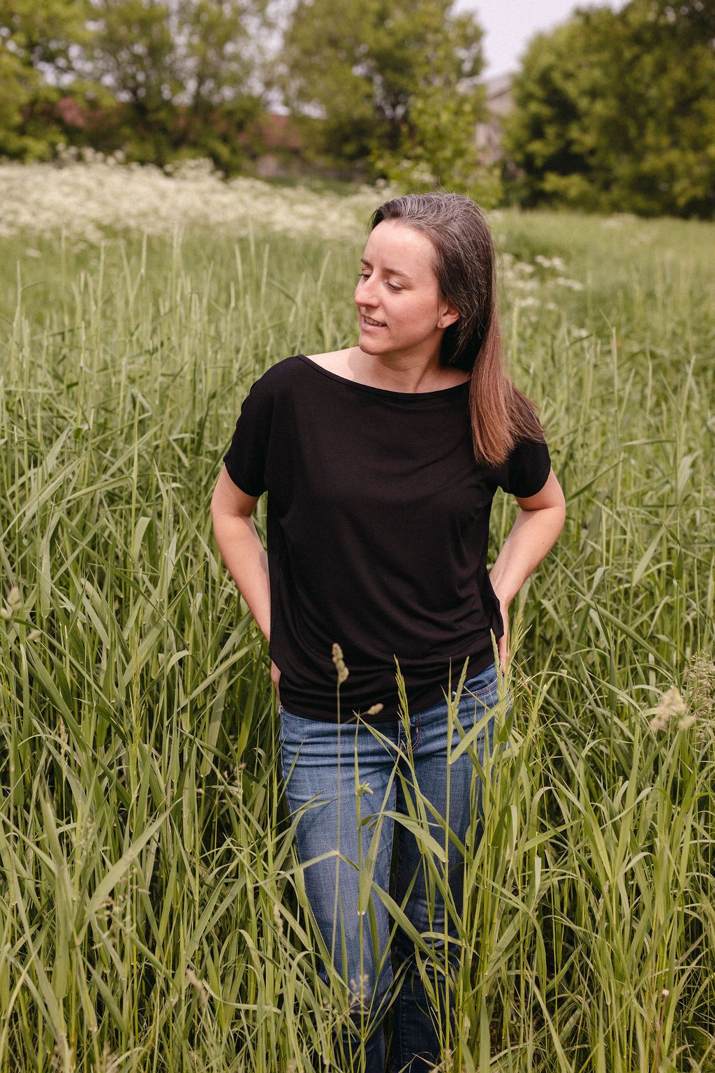 Woman standing in a field of tall green grass with trees in the background wearing a black top