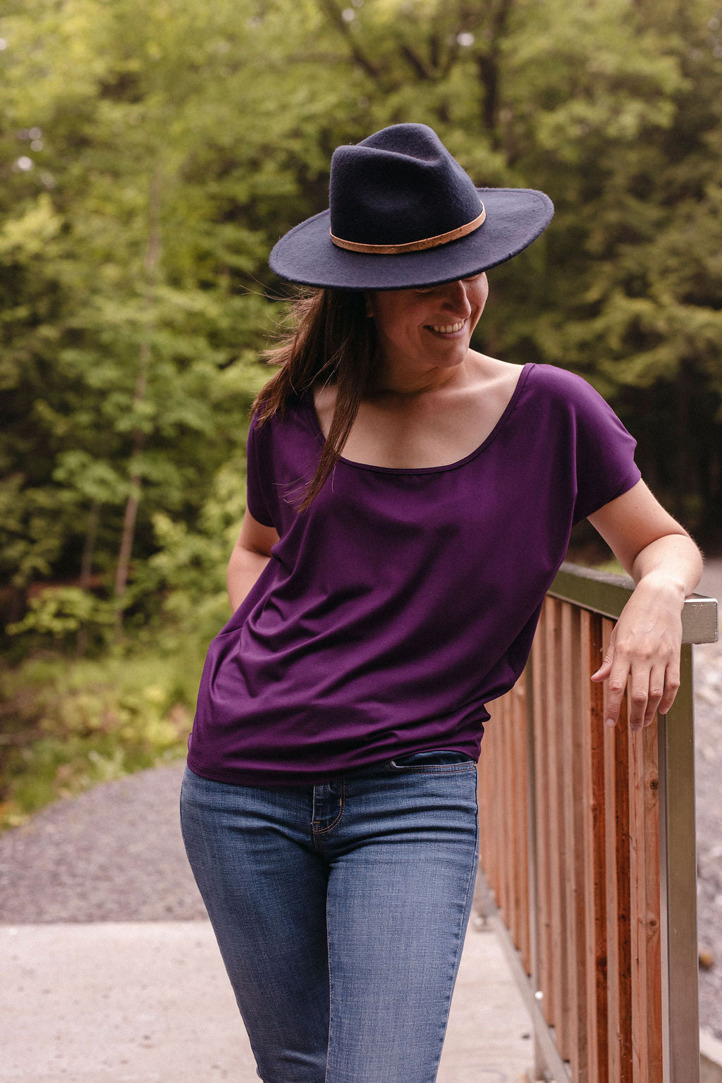 Woman wearing a purple shirt and blue jeans with a navy hat, standing outdoors.