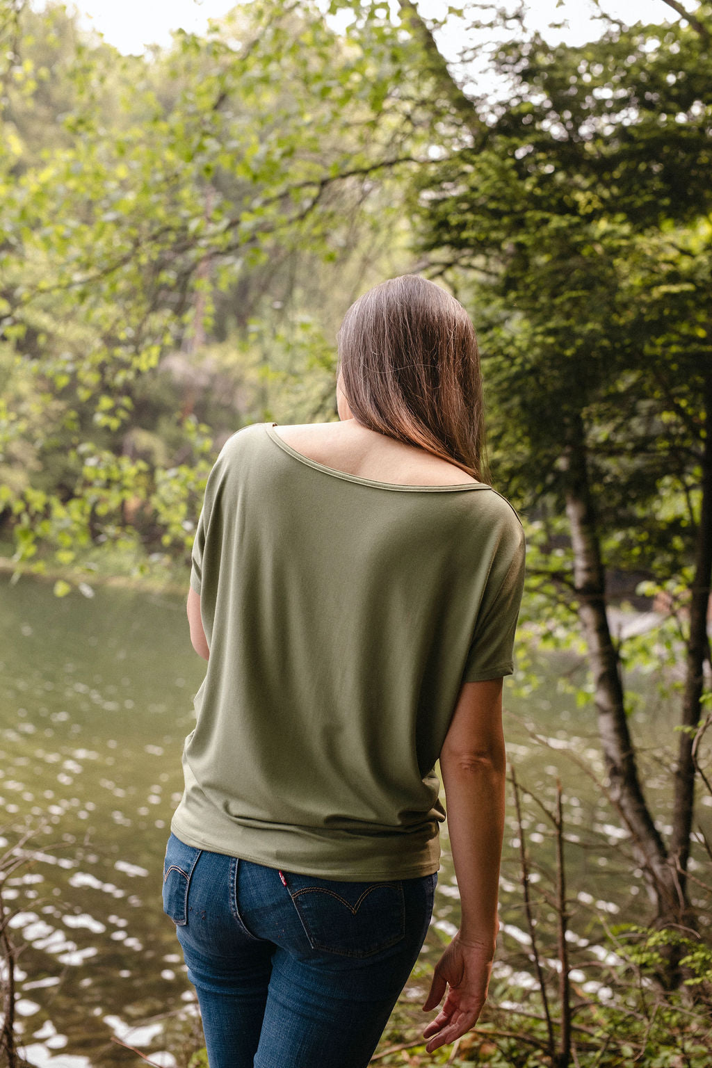 A woman standing by a lake, viewed from behind, wearing a sage green top with trees in the background