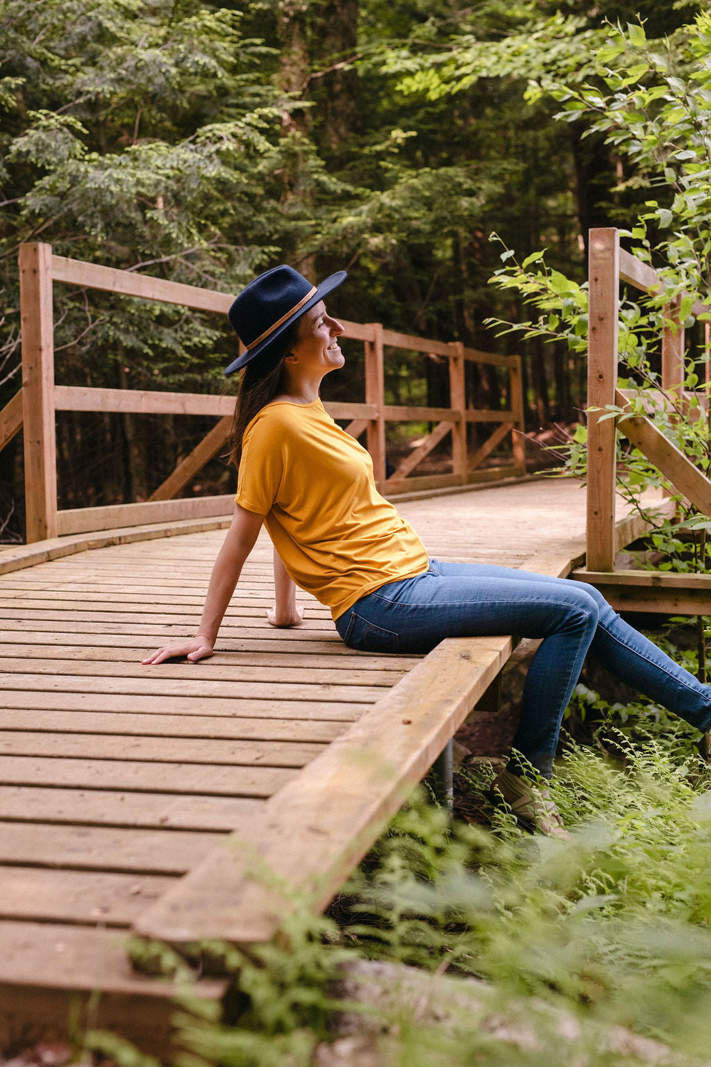 A woman sitting on a wooden bridge in a forest, wearing a yellow top, blue jeans and a navy hat