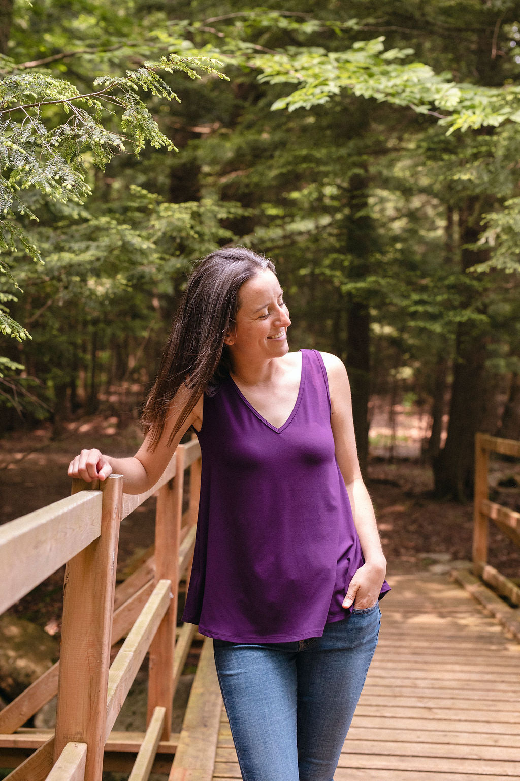 A woman wearing a purple sleeveless V-neck top, standing on a wooden bridge in a forest