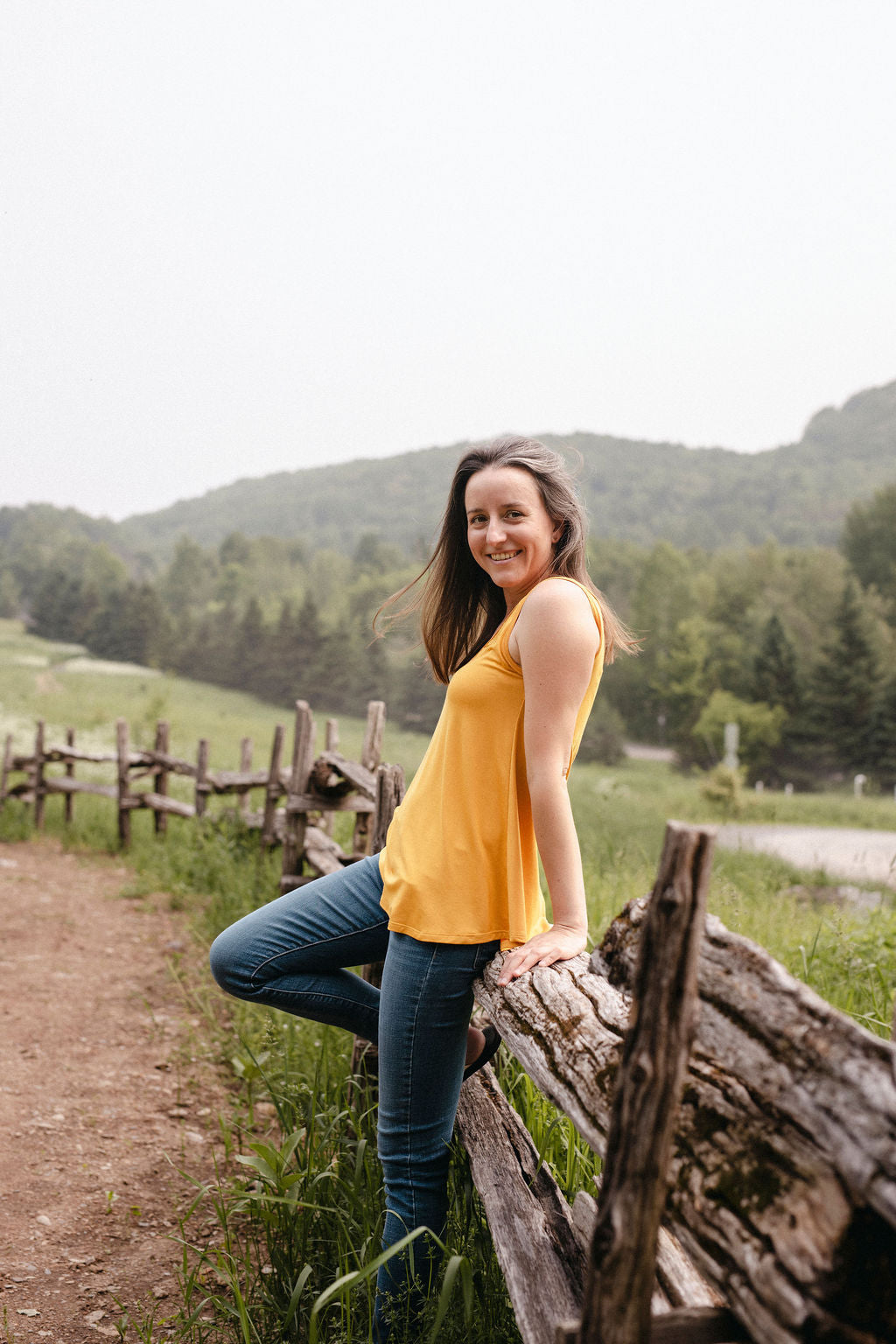 Woman in a yellow top and blue jeans sitting on a wooden fence with a scenic background of mountains and trees.