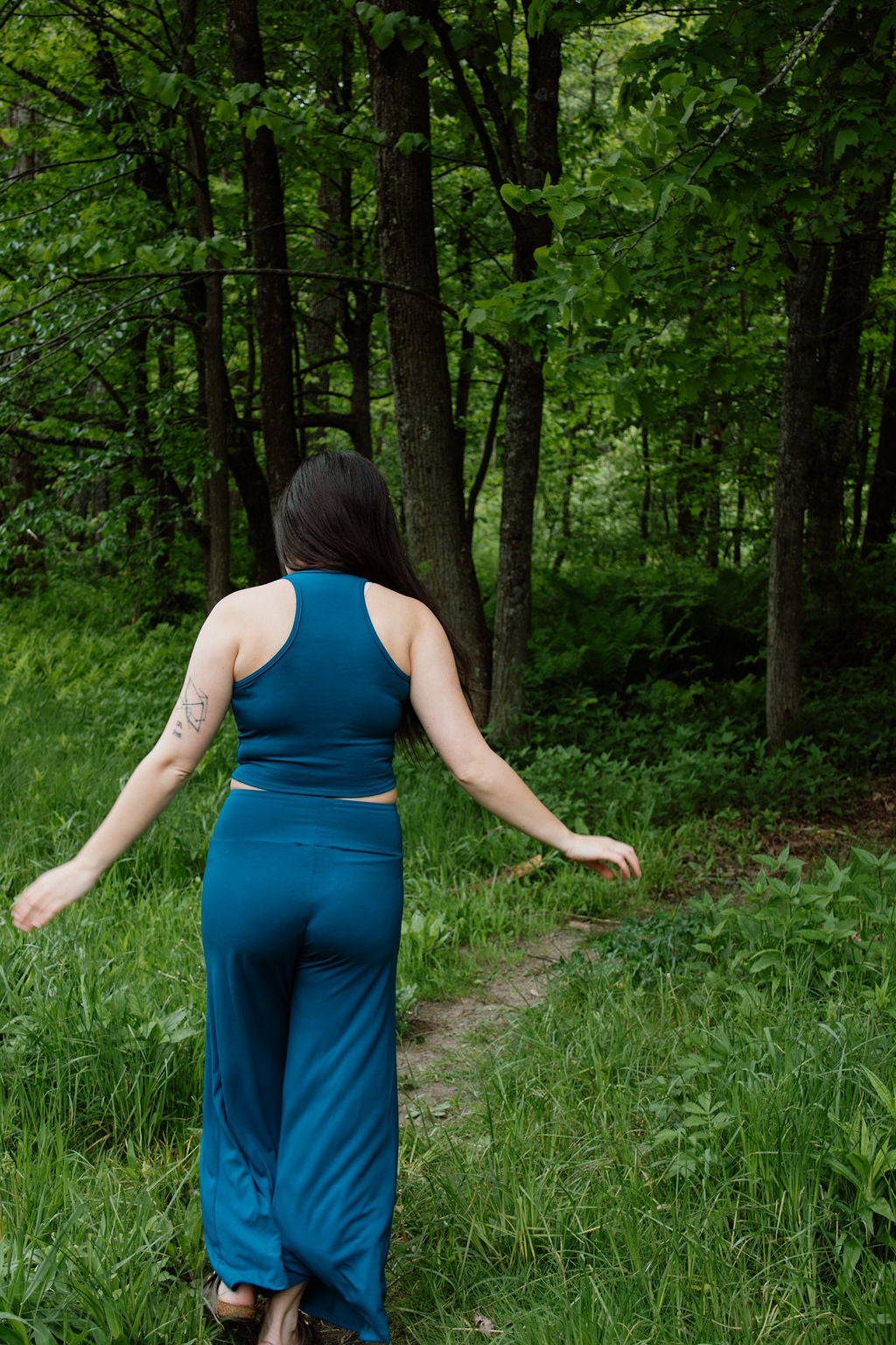 Person in a teal crop top and palazzo pants walking away from through a forest