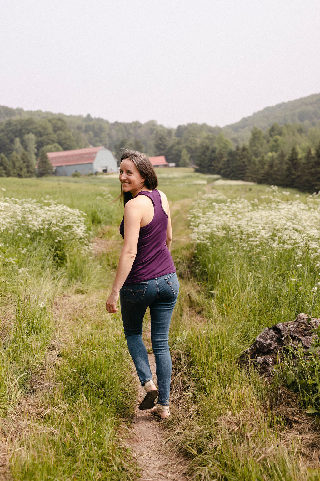 A woman wearing a plum racerback tank top, walking on a path through a field with a barn and trees in the background