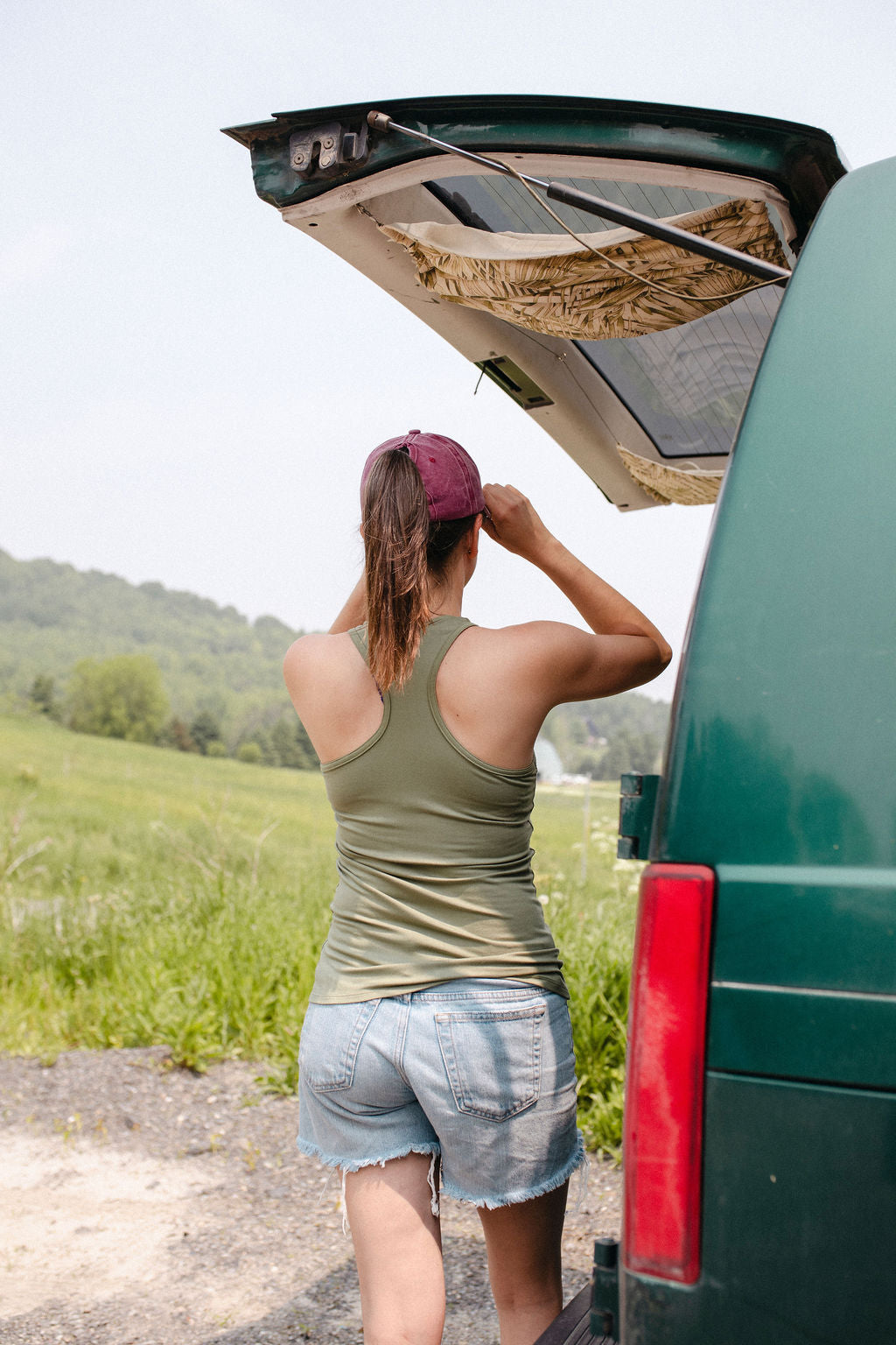 A woman viewed from behind wearing a sage green racerback tank top, standing next to an open car trunk in a natural setting