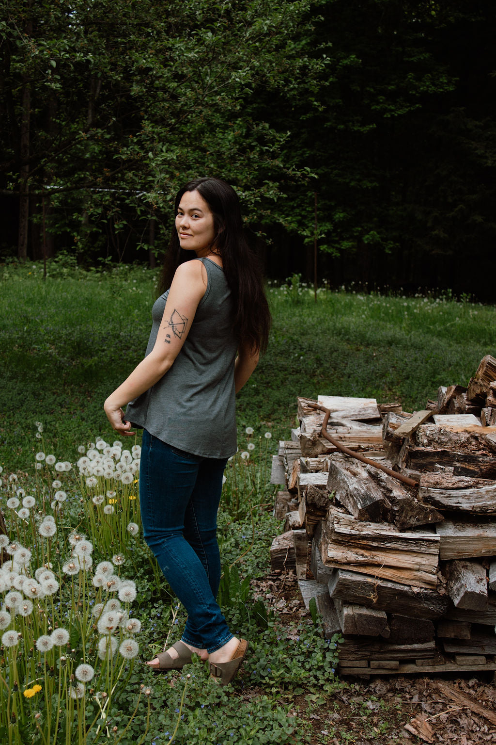 Woman standing in a natural setting with a stack of firewood and greenery.