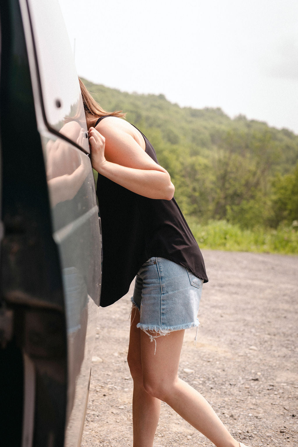 Person with black tanp top and blue jeans short leaning against a vehicle on a scenic road with greenery in the background