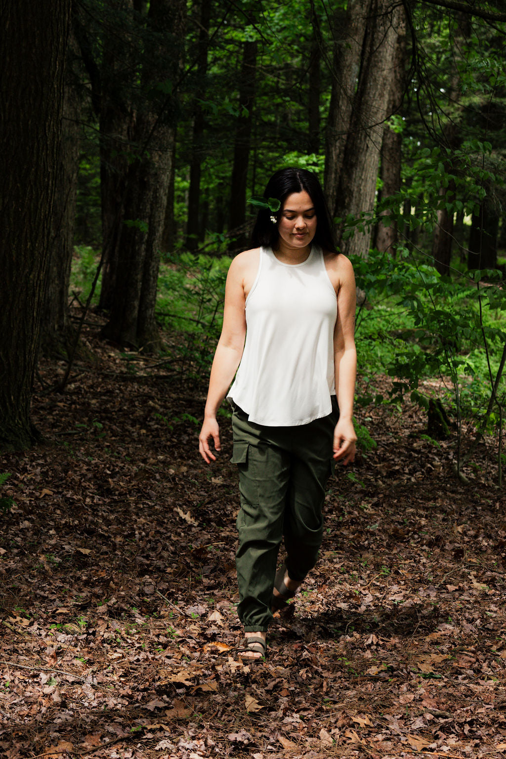 Person walking through a forest wearing a white sleeveless top and green pants.