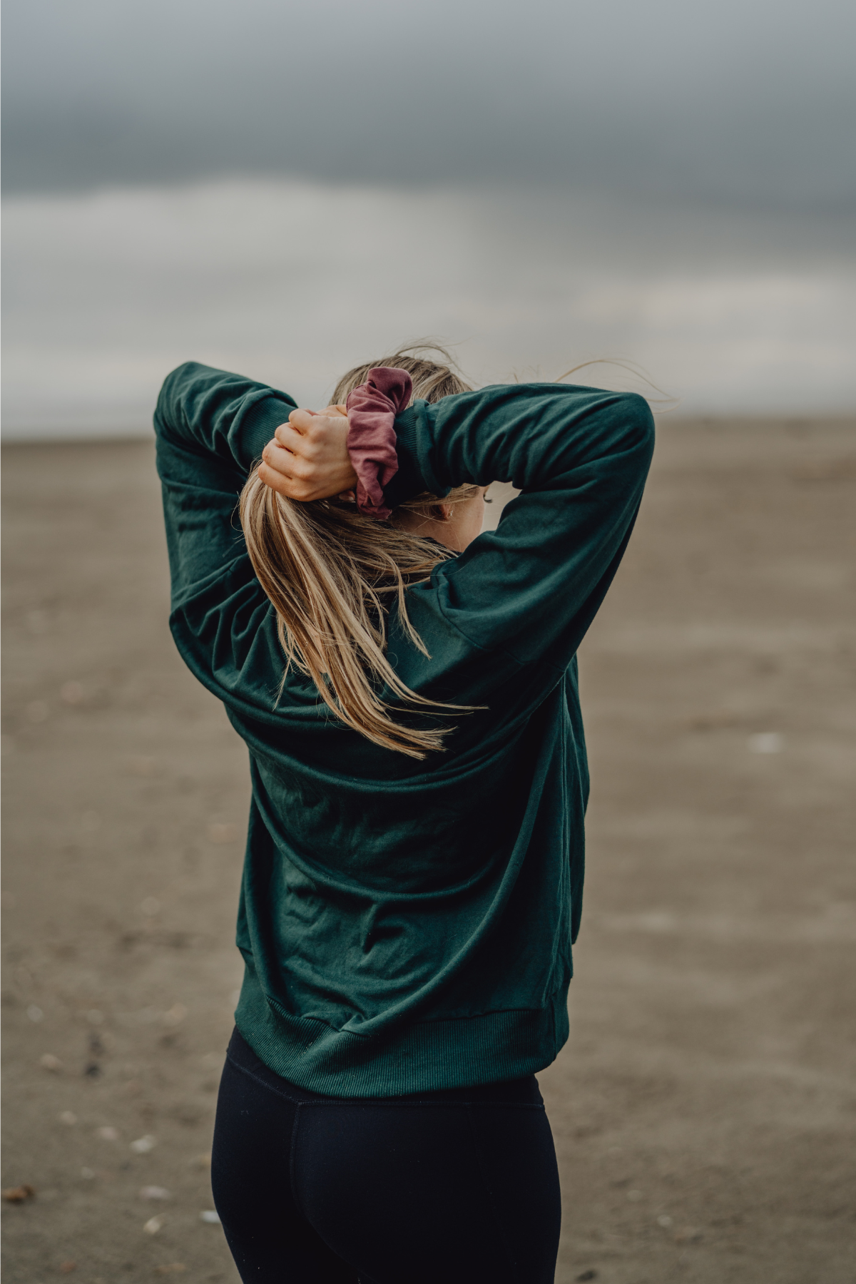 A woman viewed from behind tying their hair into a ponytail with a pink French terry scrunchie, with the beach in the background
