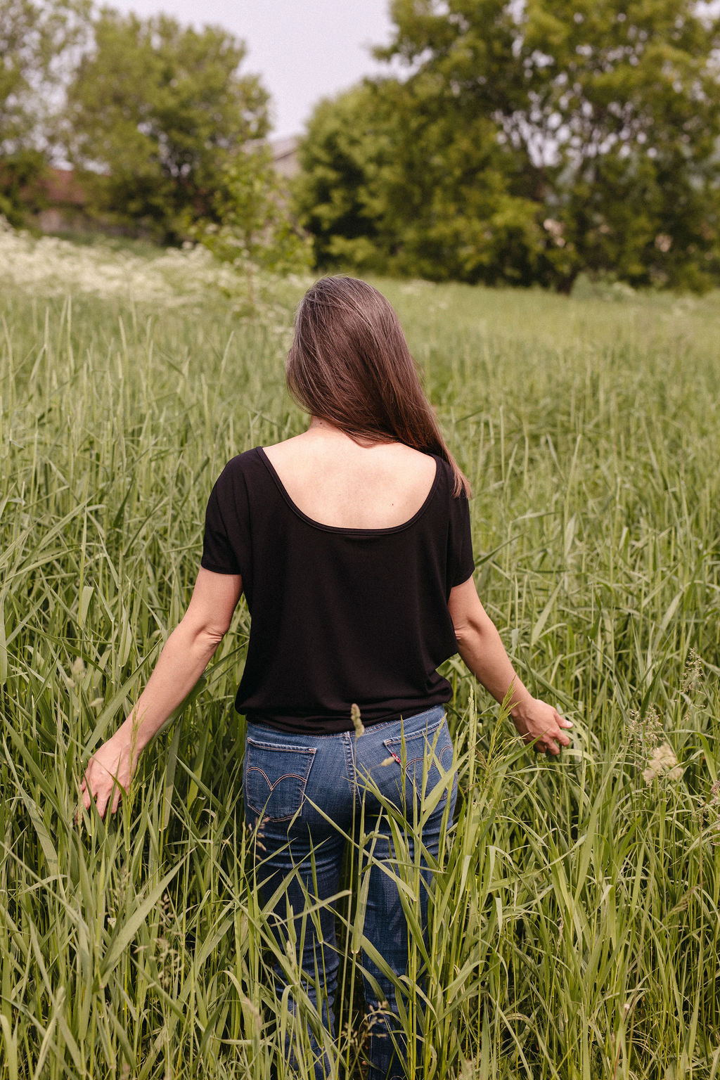 A person standing in a field of tall grass, viewed from behind, wearing a black top with trees in the background
