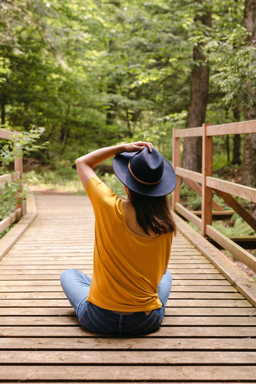 A person sitting on a wooden bridge in a forest, viewed from behind, wearing a yellow shirt, blue jeans and a navy hat