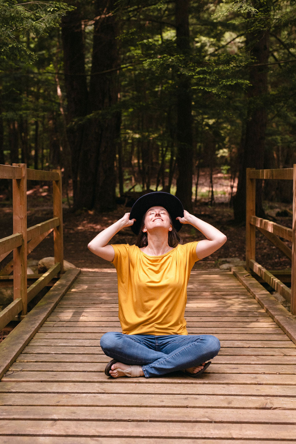 Person sitting on a wooden bridge in a forest, wearing a yellow shirt and blue jeans.