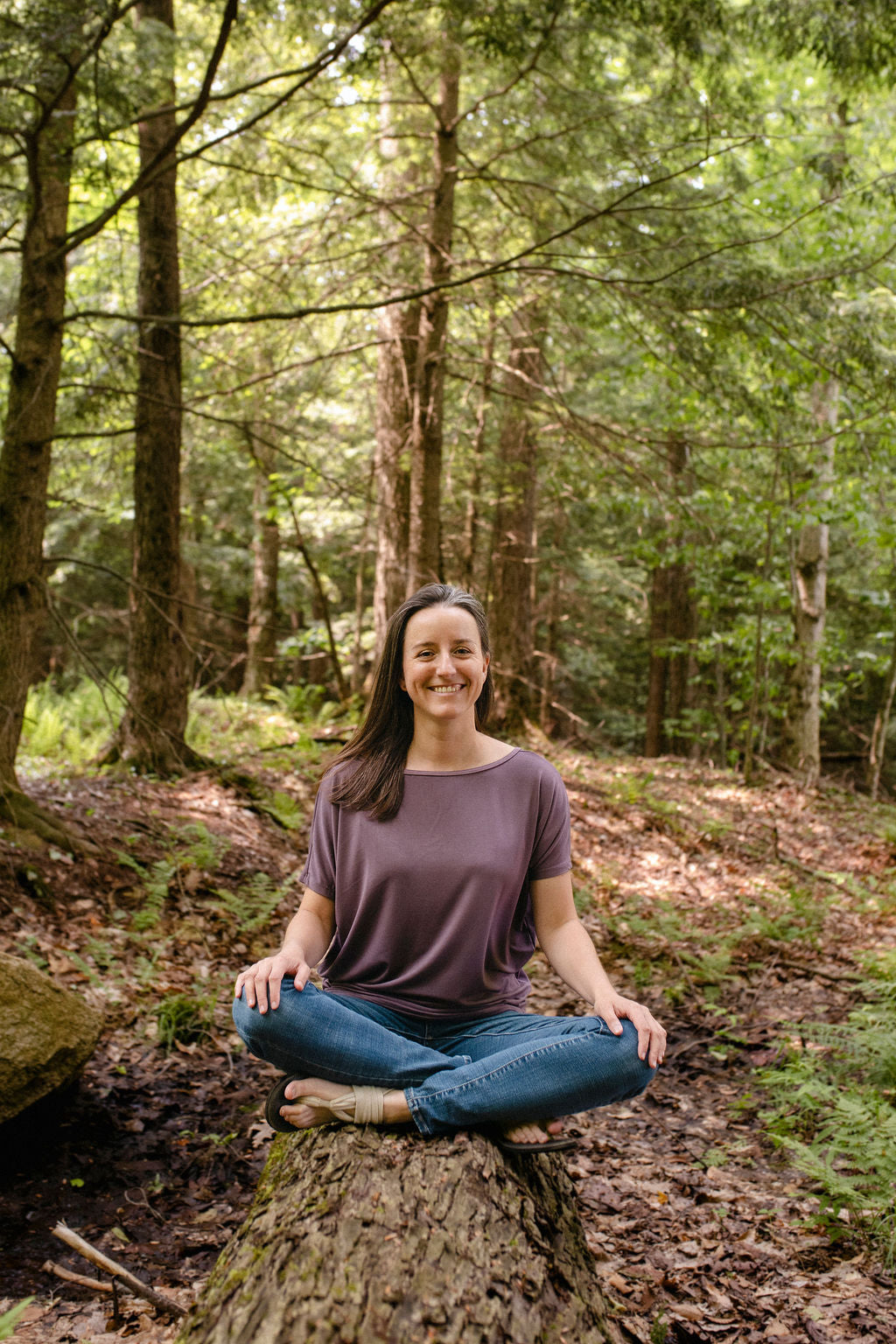 Woman sitting cross-legged on a log in a forest wearing a lavender top
