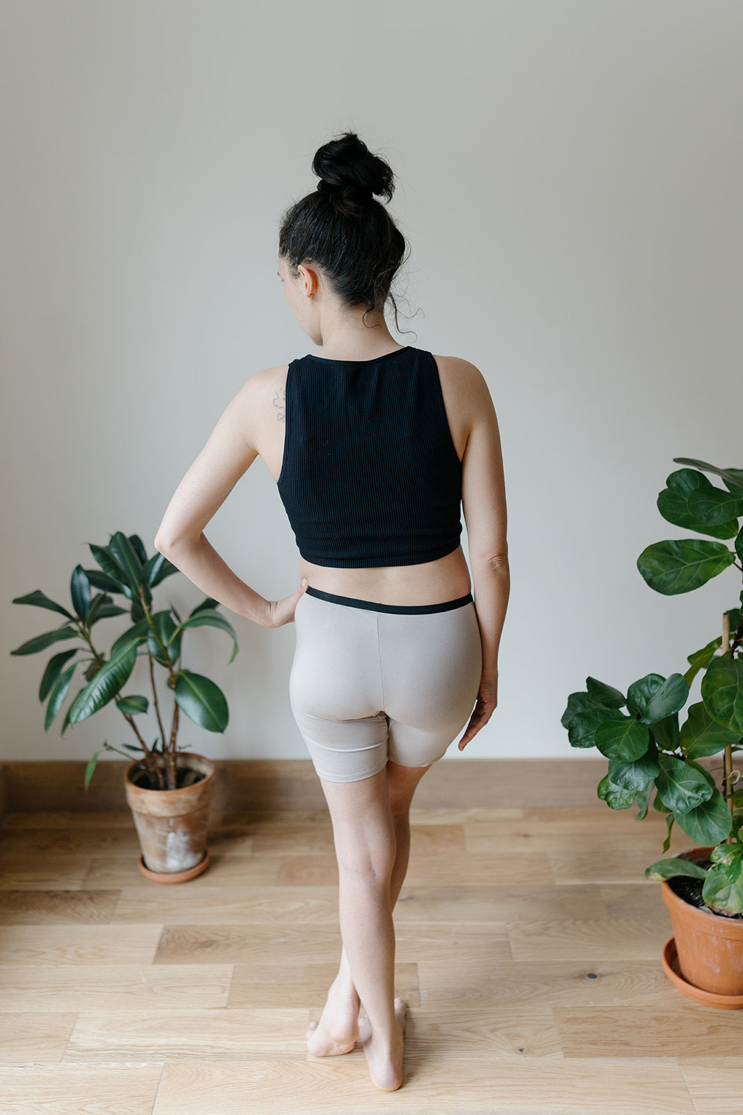A woman wearing a black top and taupe long bamboo boxer shorts, standing with her back facing the camera in a room with plants in the background