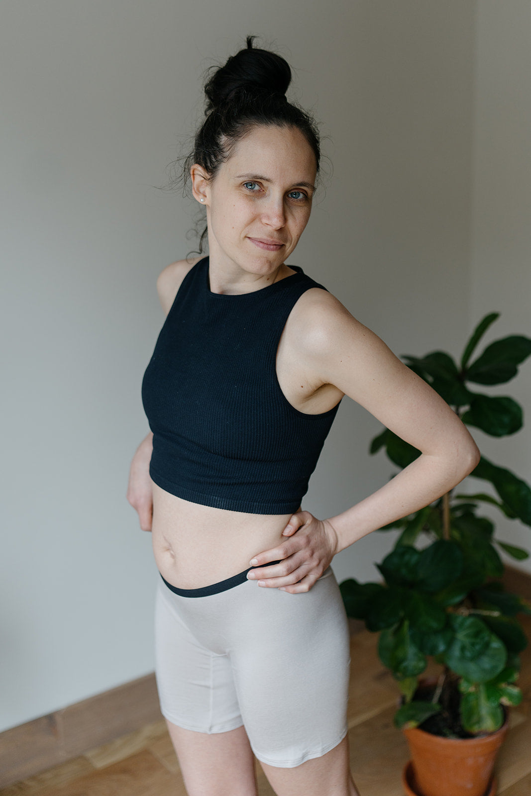 A woman wearing a black top and taupe long bamboo boxer shorts, standing in a room with plants in the background