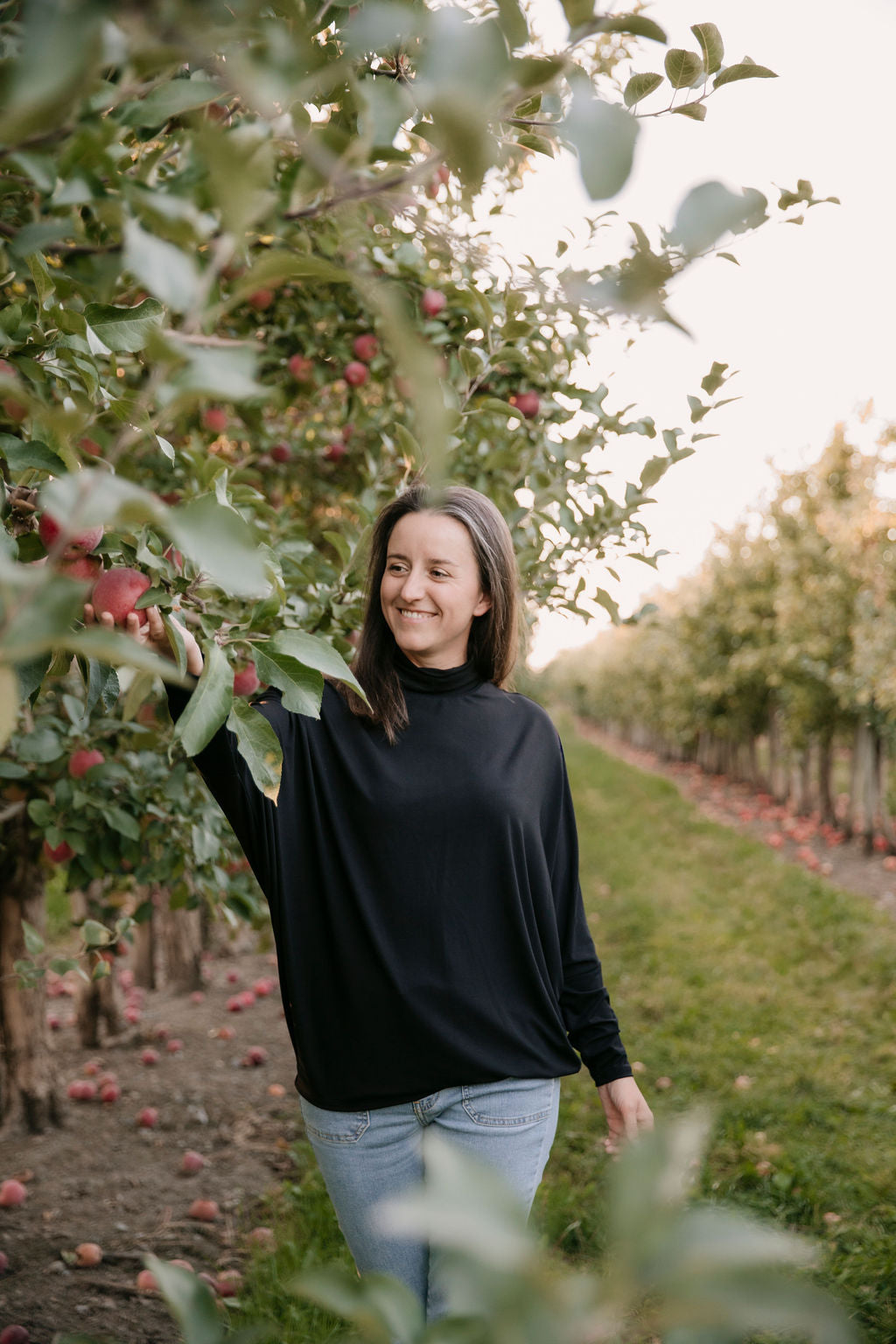 Woman wearing the Porto Batwing Long-Sleeve Top in Black and jeans, picking an apple in an orchard with apples on the trees.