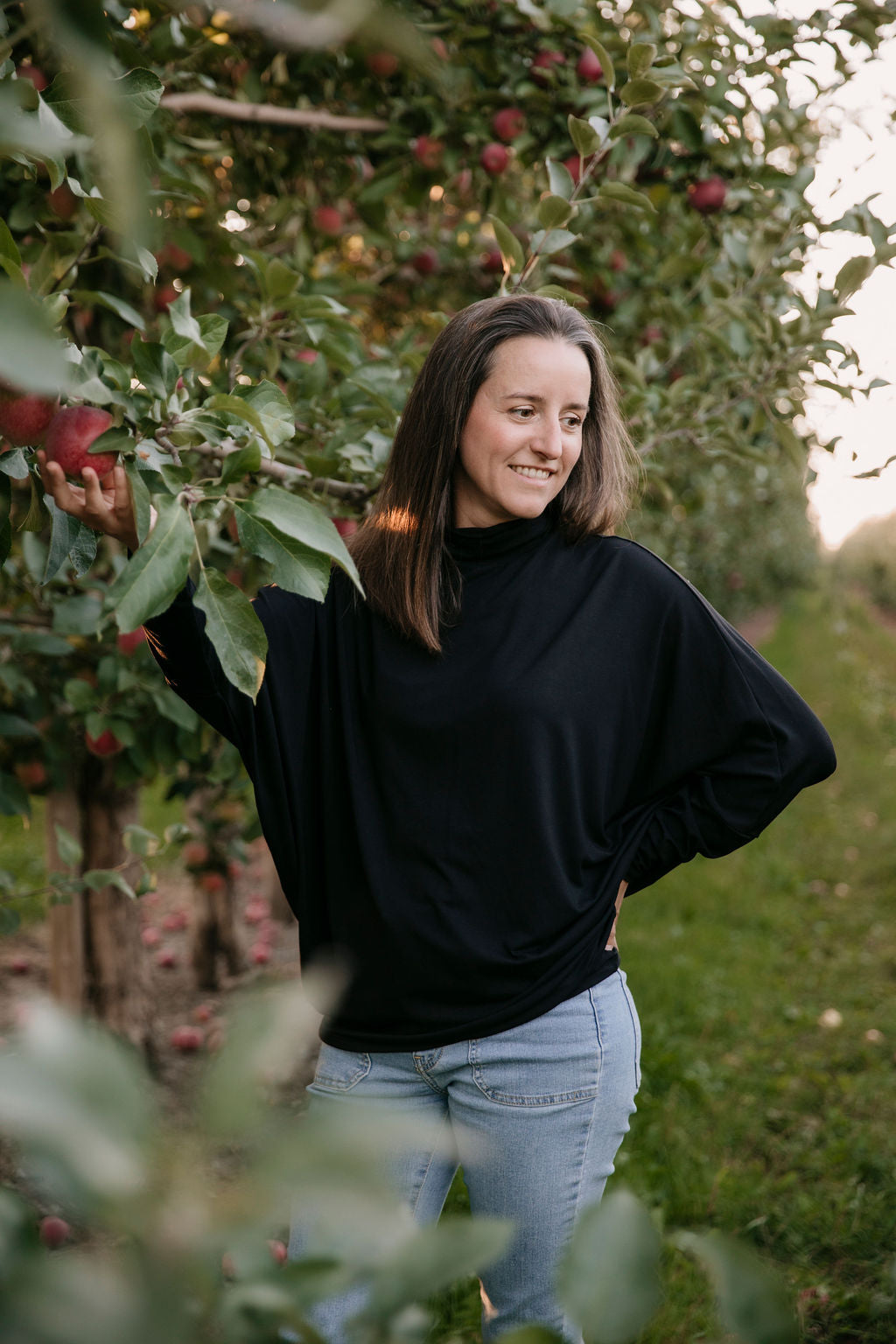 Woman wearing the Porto Batwing Long-Sleeve Top in Black and jeans, standing in an apple orchard with apples on the trees.