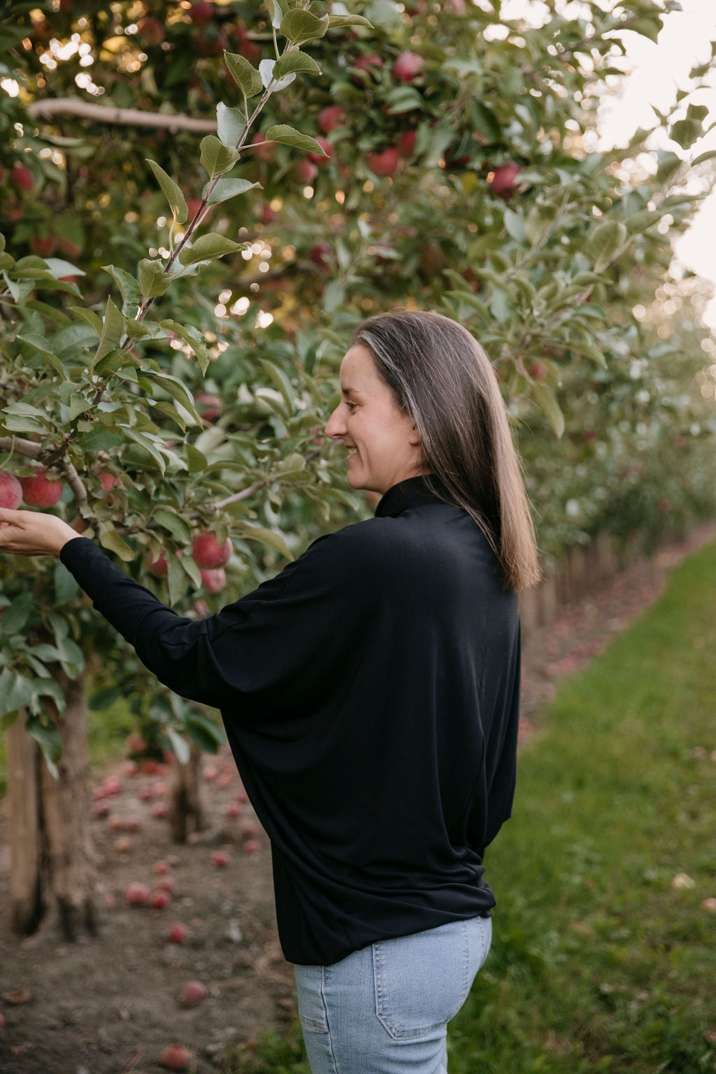 Woman wearing the Porto Batwing Long-Sleeve Top in Black and jeans, picking an apple in an orchard with apples on the trees.