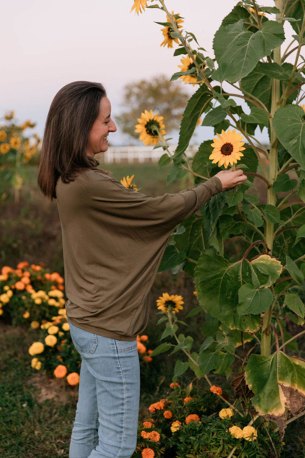 Woman wearing The Batwing Long-Sleeve Top in Olive, touching a tall sunflower in a garden.
