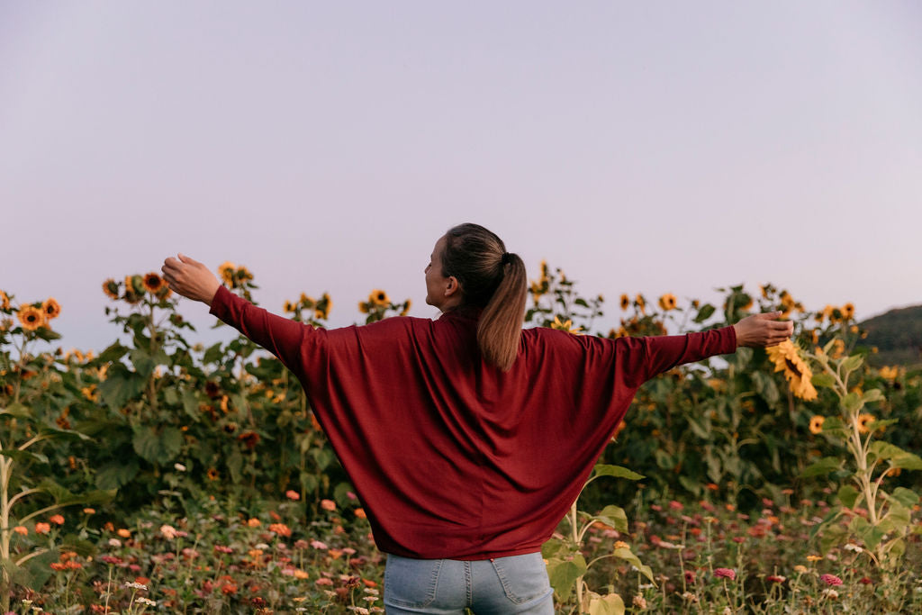 Woman from behind wearing the Porto Batwing Long-Sleeve Top standing in a sunflower field with her arms spread open.