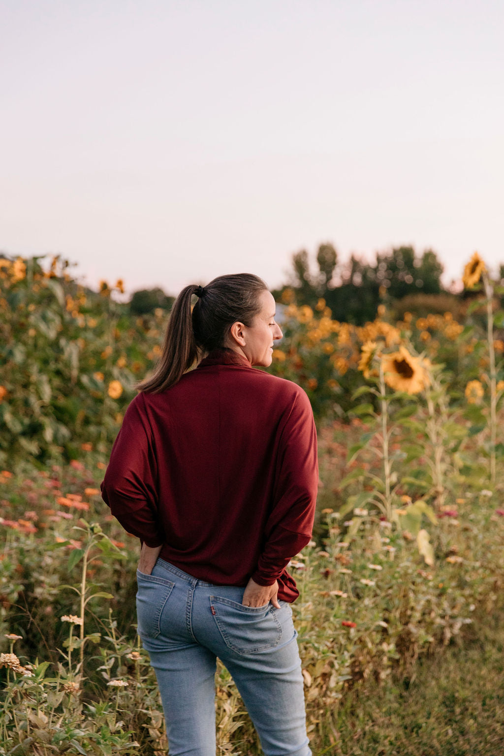 Woman from behind wearing the Porto Batwing Long-Sleeve Top and blue jeans standing in a sunflower field.