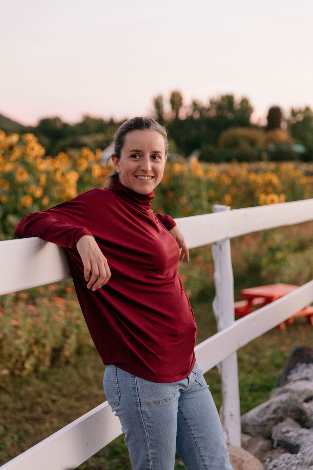Woman wearing the Porto Batwing Long-Sleeve Top standing by a white fence with a sunflower field in the background.
