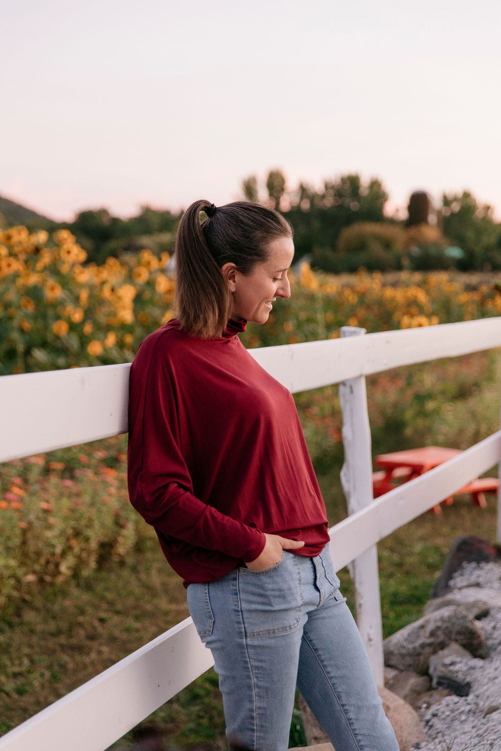 Woman wearing the Porto Batwing Long-Sleeve Top standing by a white fence with a sunflower field in the background.
