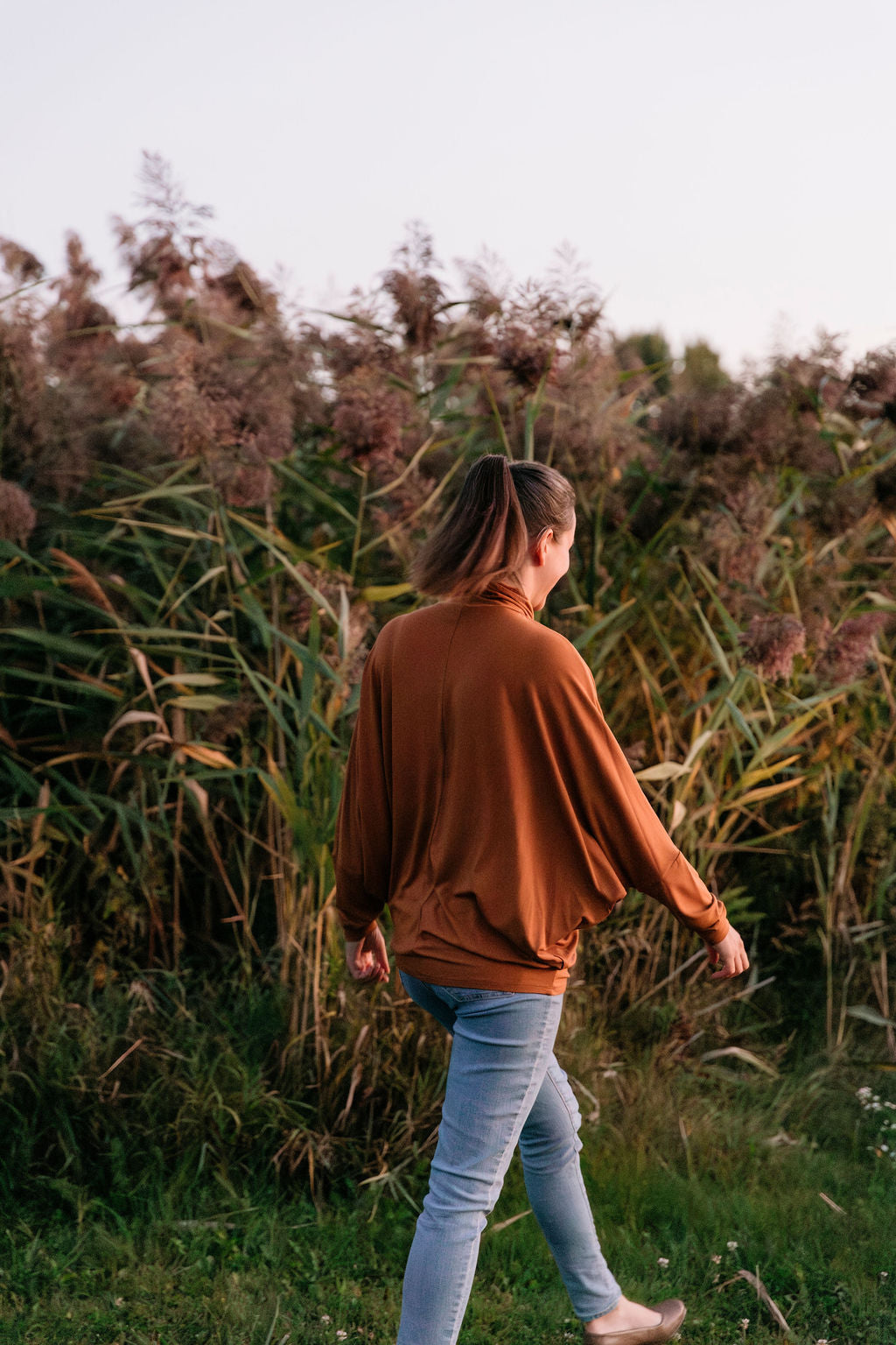 Woman wearing The Batwing Long-Sleeve Top in Redwood walking with her back to the camera in a field with tall grass.