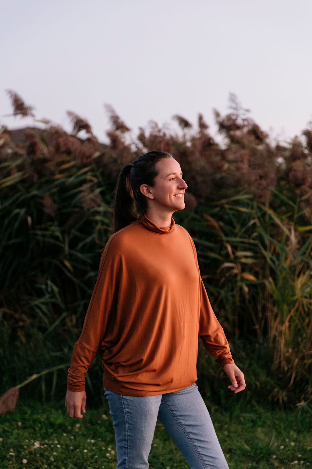 Woman wearing The Batwing Long-Sleeve Top in Redwood with blue jeans, walking in a field with tall grass.