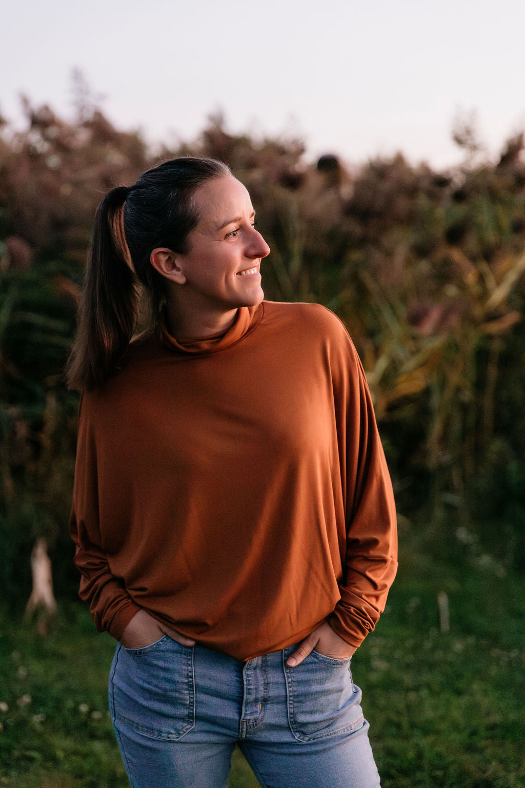Woman wearing The Batwing Long-Sleeve Top in Redwood with blue jeans, standing in a field with tall grass.