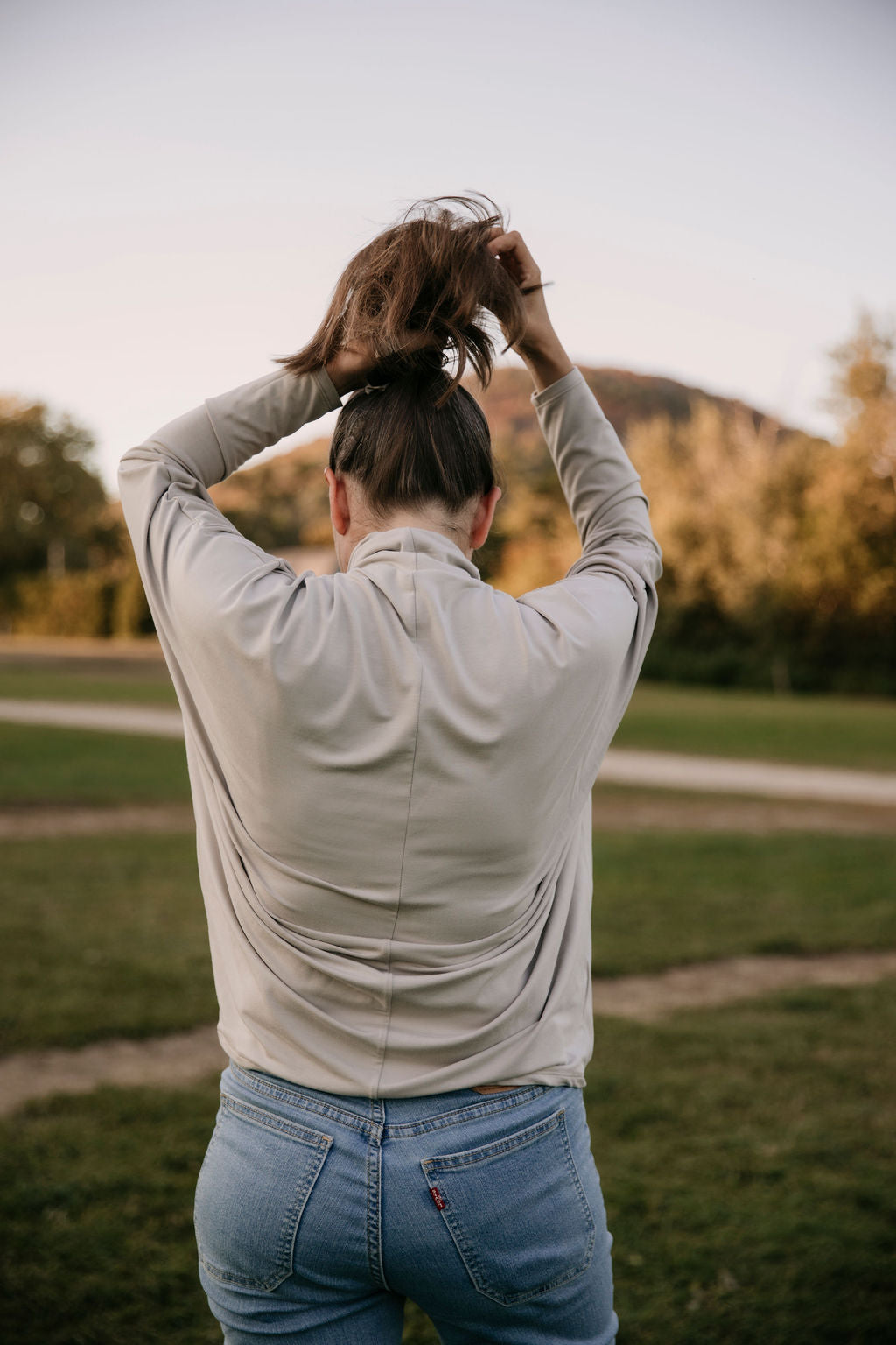 Woman wearing The Batwing Long-Sleeve Top in Taupe, standing outdoors with trees in the background, seen from behind, her arms up making a ponytail.