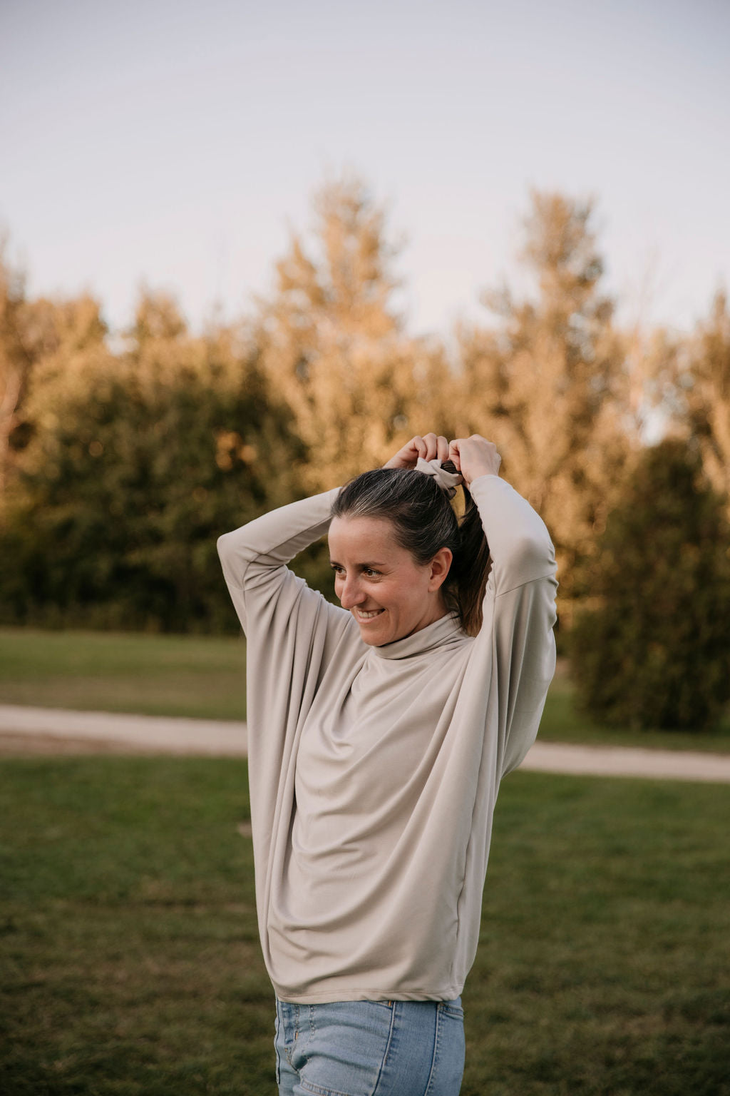 Woman wearing The Batwing Long-Sleeve Top in Taupe, standing outdoors with trees in the background her arms up making a ponytail.