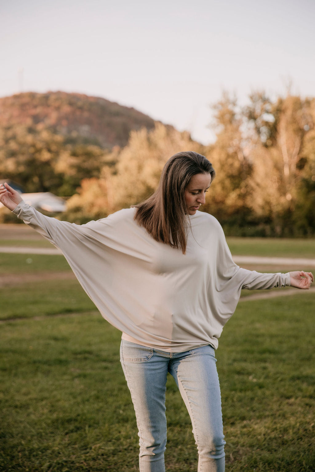 Woman wearing The Batwing Long-Sleeve Top in Taupe, standing outdoors with trees in the background, her arms swinging in the air.