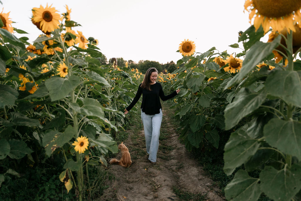 Woman wearing The Breezy Long-Sleeve Shirt in Black, walking in a sunflower field next to an orange cat.