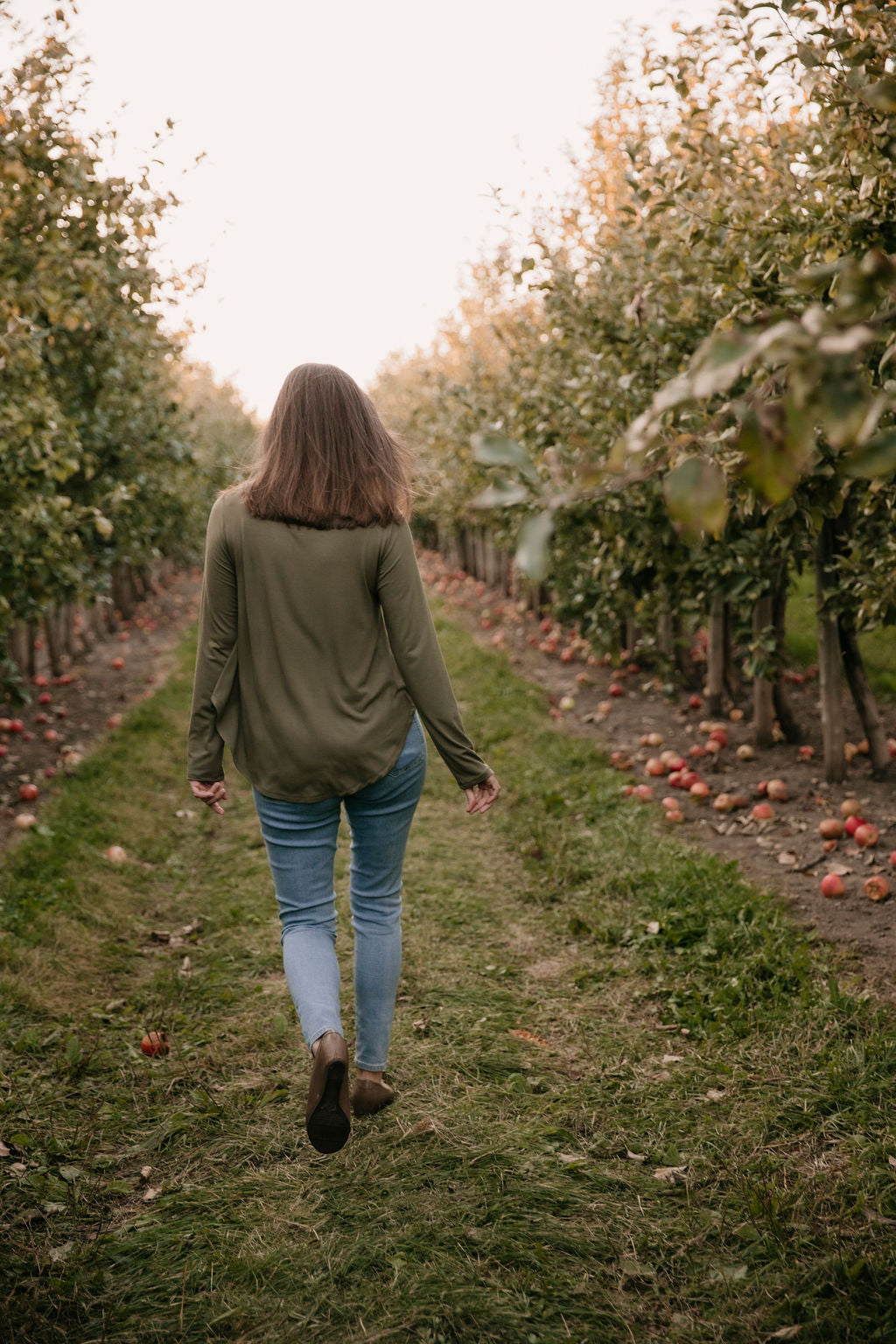 Woman wearing The Breezy Long-Sleeve Shirt in Olive, walking in an orchard, seen from behind.