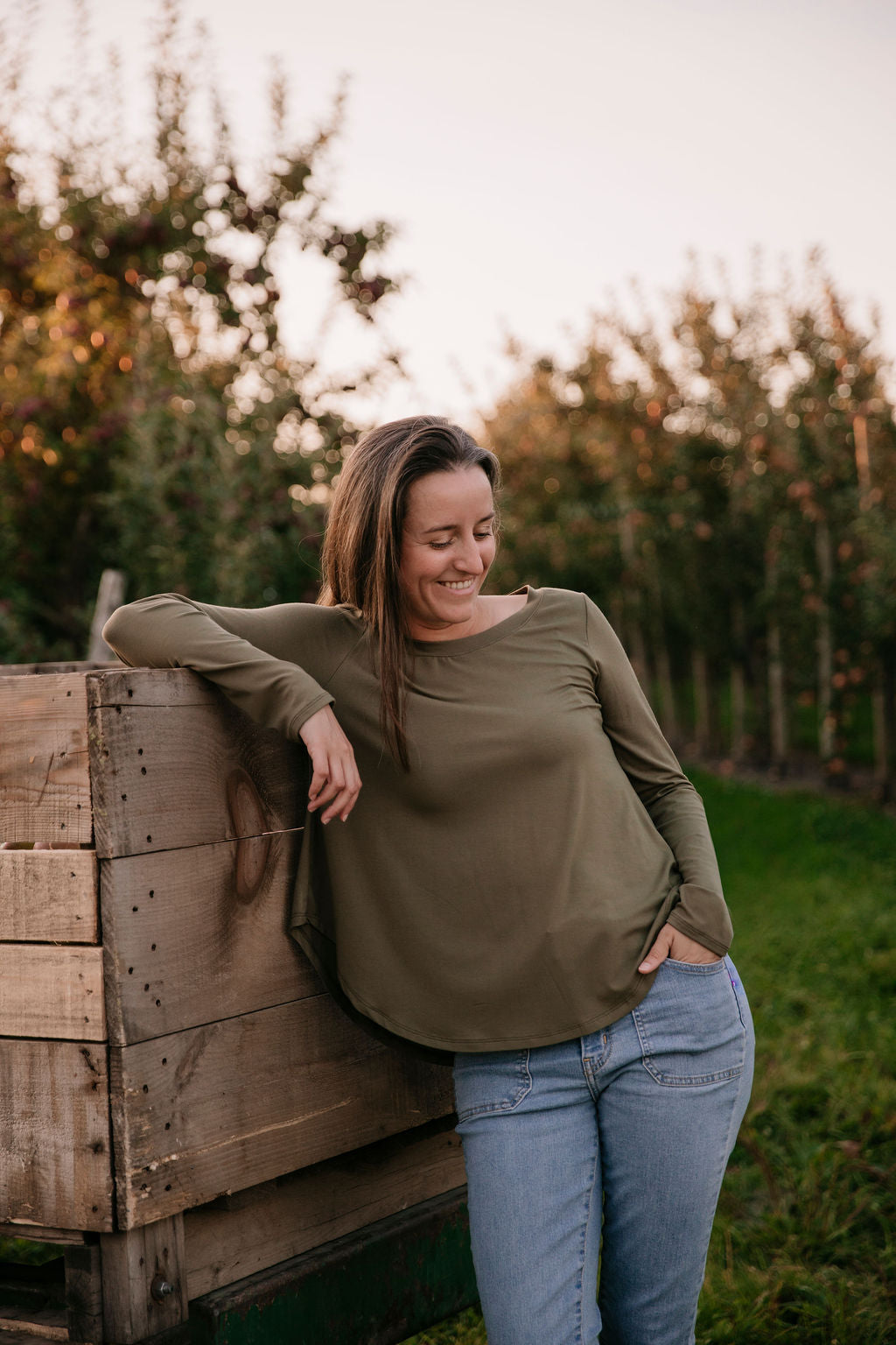 Woman wearing The Breezy Long-Sleeve Shirt in Olive, standing in front of wooden crates with a sunset and an orchard in the background.