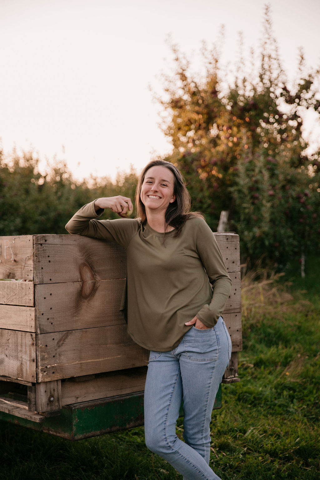 Woman wearing The Breezy Long-Sleeve Shirt in Olive, standing in front of wooden crates with a sunset and an orchard in the background.
