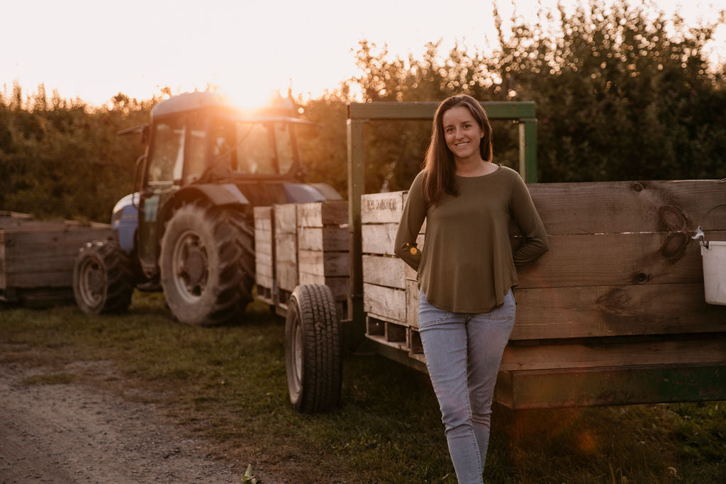 Woman wearing The Breezy Long-Sleeve Shirt in Olive, standing in front of wooden crates and a tractor, with a sunset in the background.