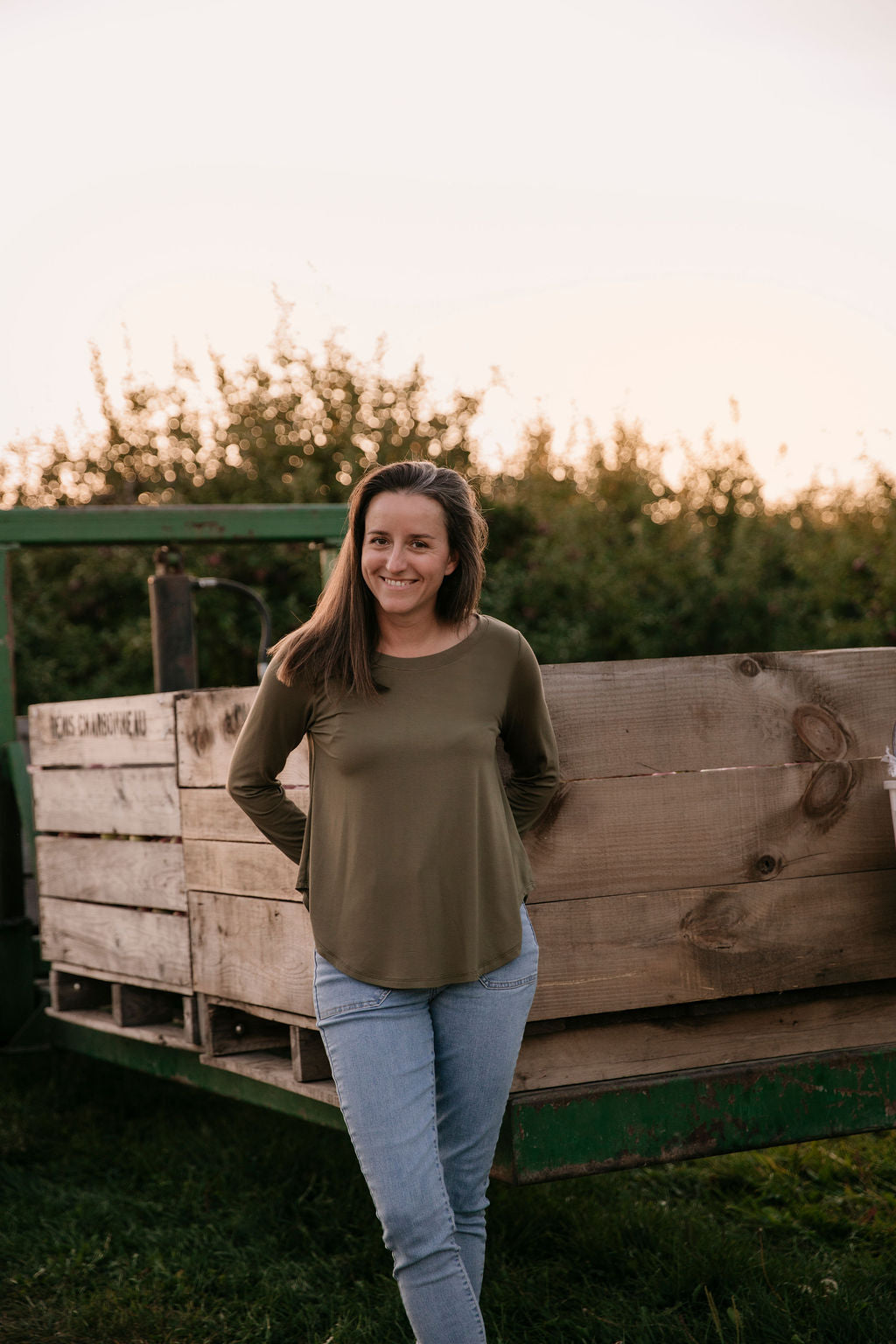 Woman wearing The Breezy Long-Sleeve Shirt in Olive, standing in front of wooden crates with a sunset in the background.