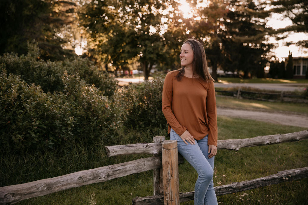 Woman wearing The Breezy Long-Sleeve Shirt in Redwood and light blue jeans, sitting on a wooden fence.