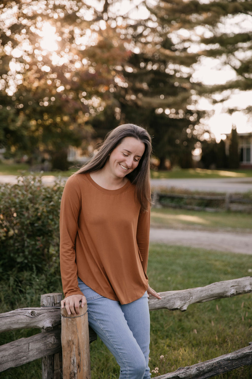 Woman wearing The Breezy Long-Sleeve Shirt in Redwood and light blue jeans, sitting on a wooden fence.