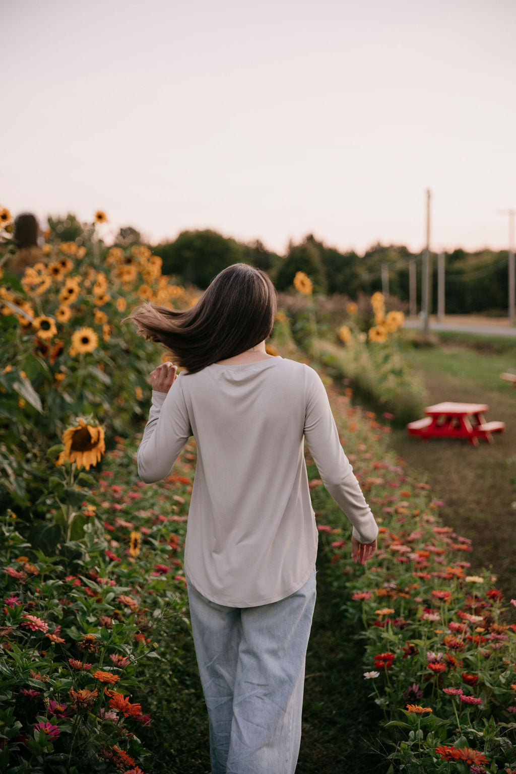Woman wearing The Breezy Long-Sleeve Shirt in Taupe, walking in a field of sunflowers and other flowers, seen from behind, flicking her hair.
